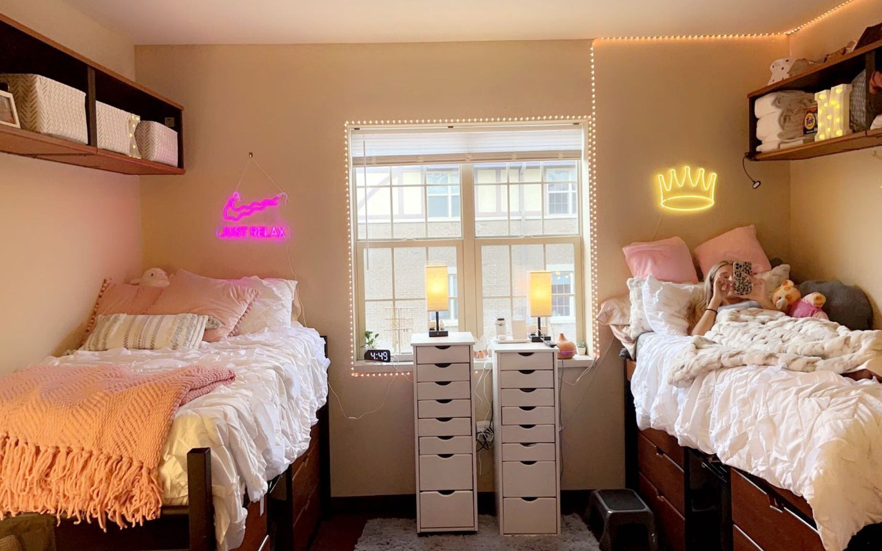 Room decorated with white lights and beds with white and pink linens. A female student is reading her phone on the bed to the right