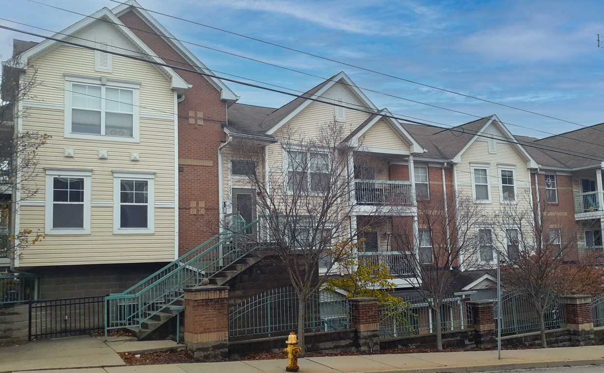 Exterior of Bellevue Gardens. There are balconies and window visible on the apartment building. There is a green iron gate with brick posts in front of the building and a row of trees in front of the gate.