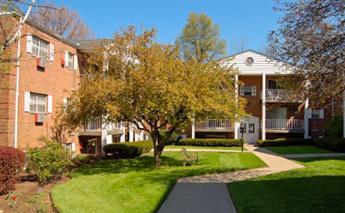 Exterior of Senator Place. There is a sidewalk with green grass on either side that path goes past a building on the left and directly towards a building in the background. There is a tree with a bench under it to the left of the sidewalk.